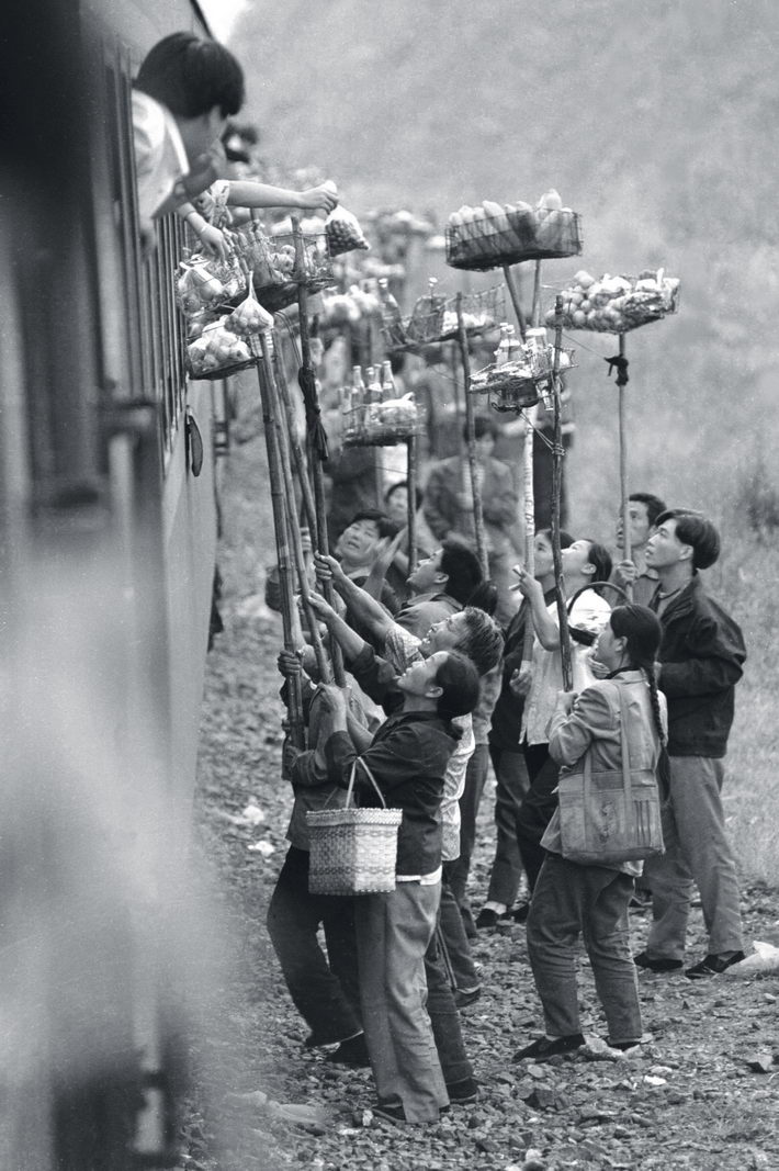 October 1996: At a small station called Qiangzilu on the railway route from Beijing to Qinhuangdao City, villagers sell local specialties to passengers. The train stopped for only five minutes and the station lacked a platform, so villagers invented a new method to sell their goods.  by Zhou Chaorong   In the mid-1990s, China was shifting from a planned economy to a market economy. Commerce prospered across the country, as evidenced by the axiom “900 million out of a billion Chinese people are businesspeople.”